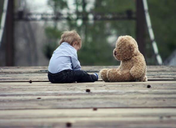 A young boy sits outside on wooden deck with a teddy bear