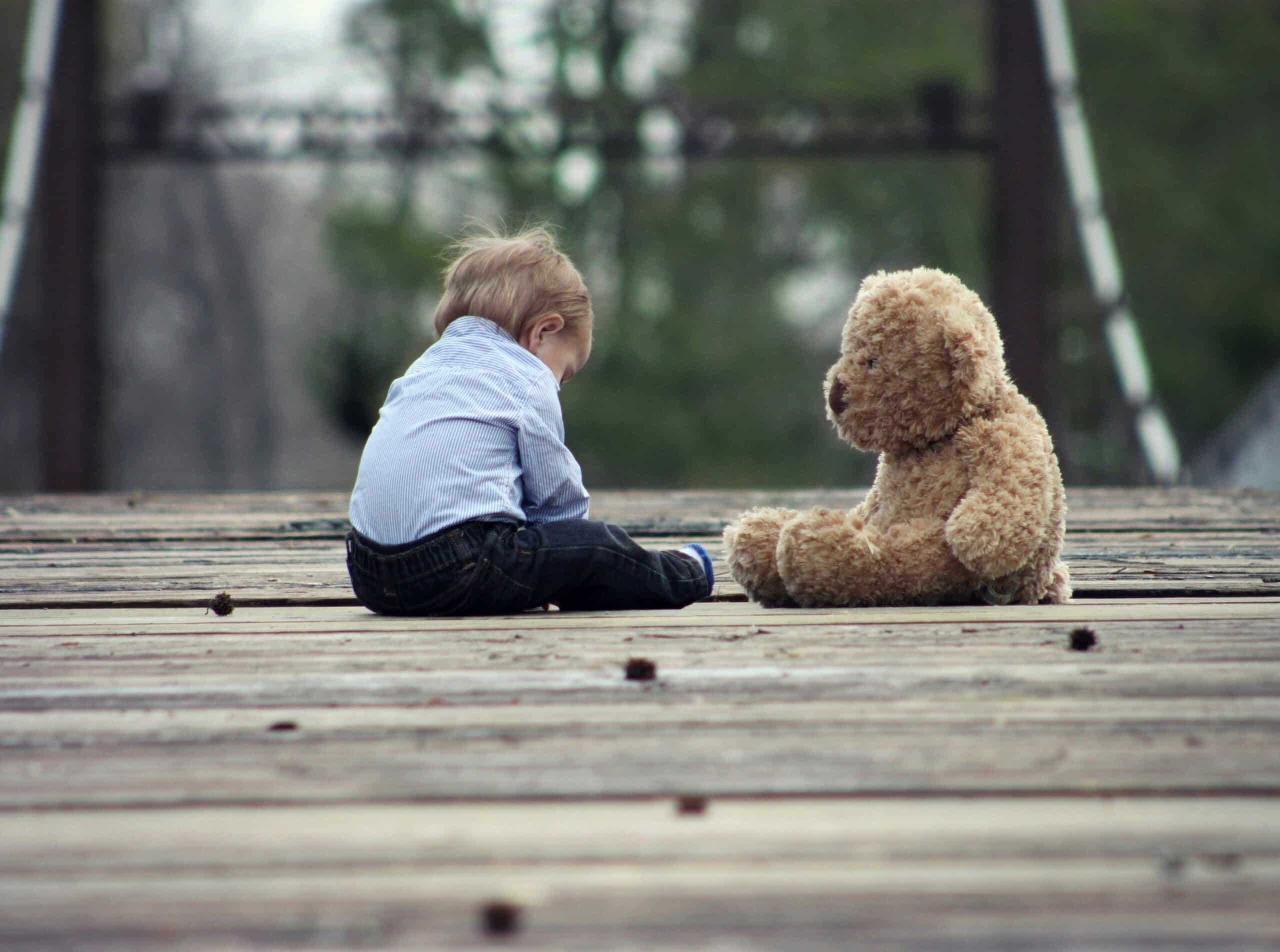 A young boy sits outside on wooden deck with a teddy bear