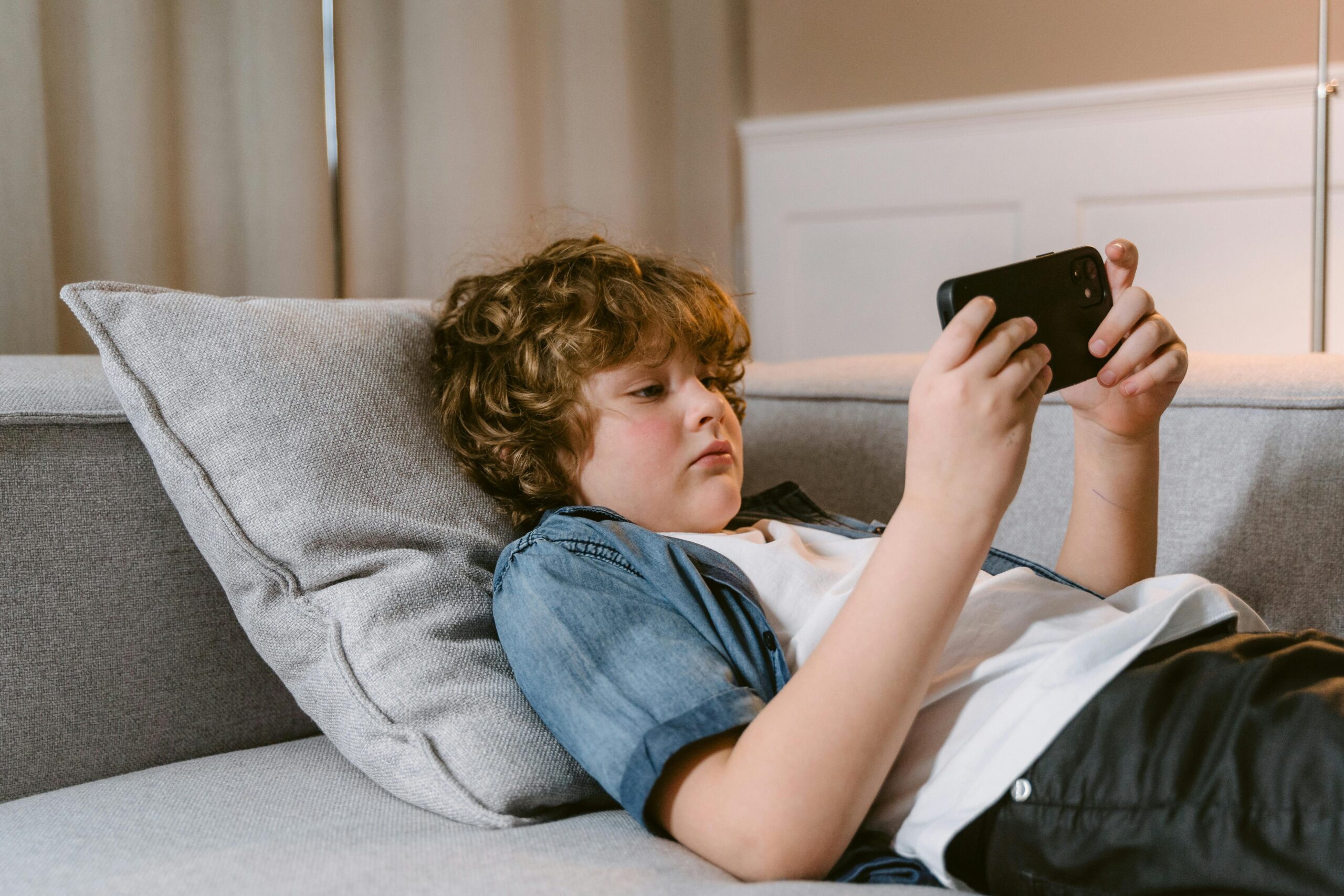 Red headed boy lying on the sofa using a mobile phone in a blue shirt