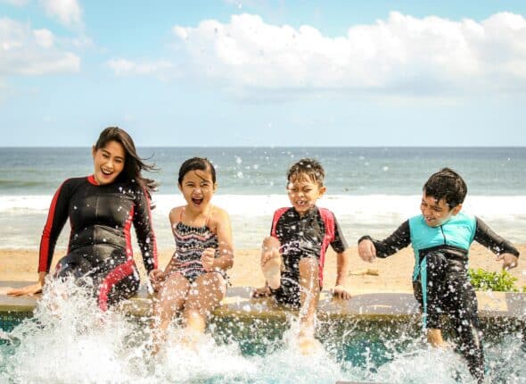 A teenage girl in a wetsuit with three kids and splashing in a sea pool at the beach.