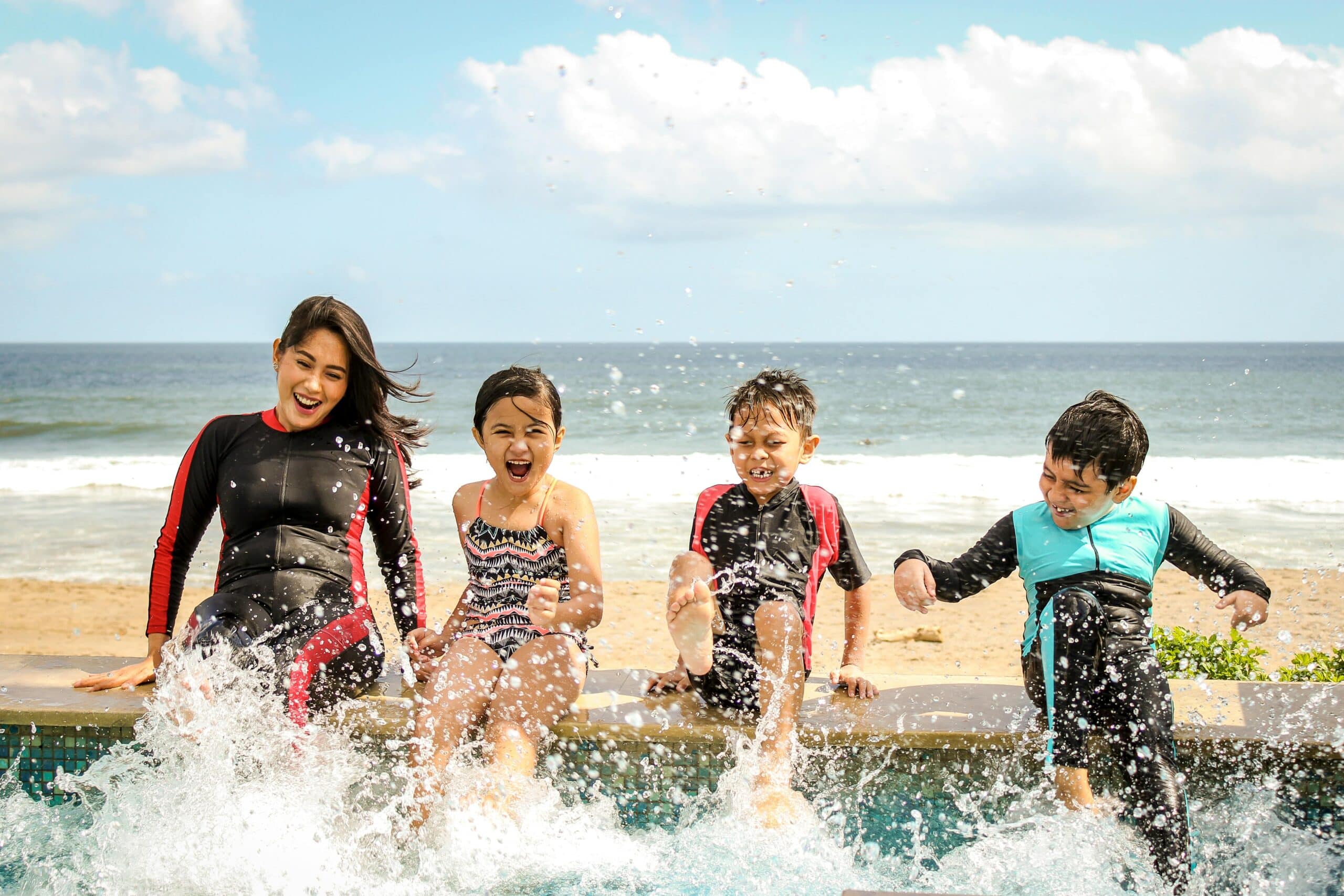 A teenage girl in a wetsuit with three kids and splashing in a sea pool at the beach.