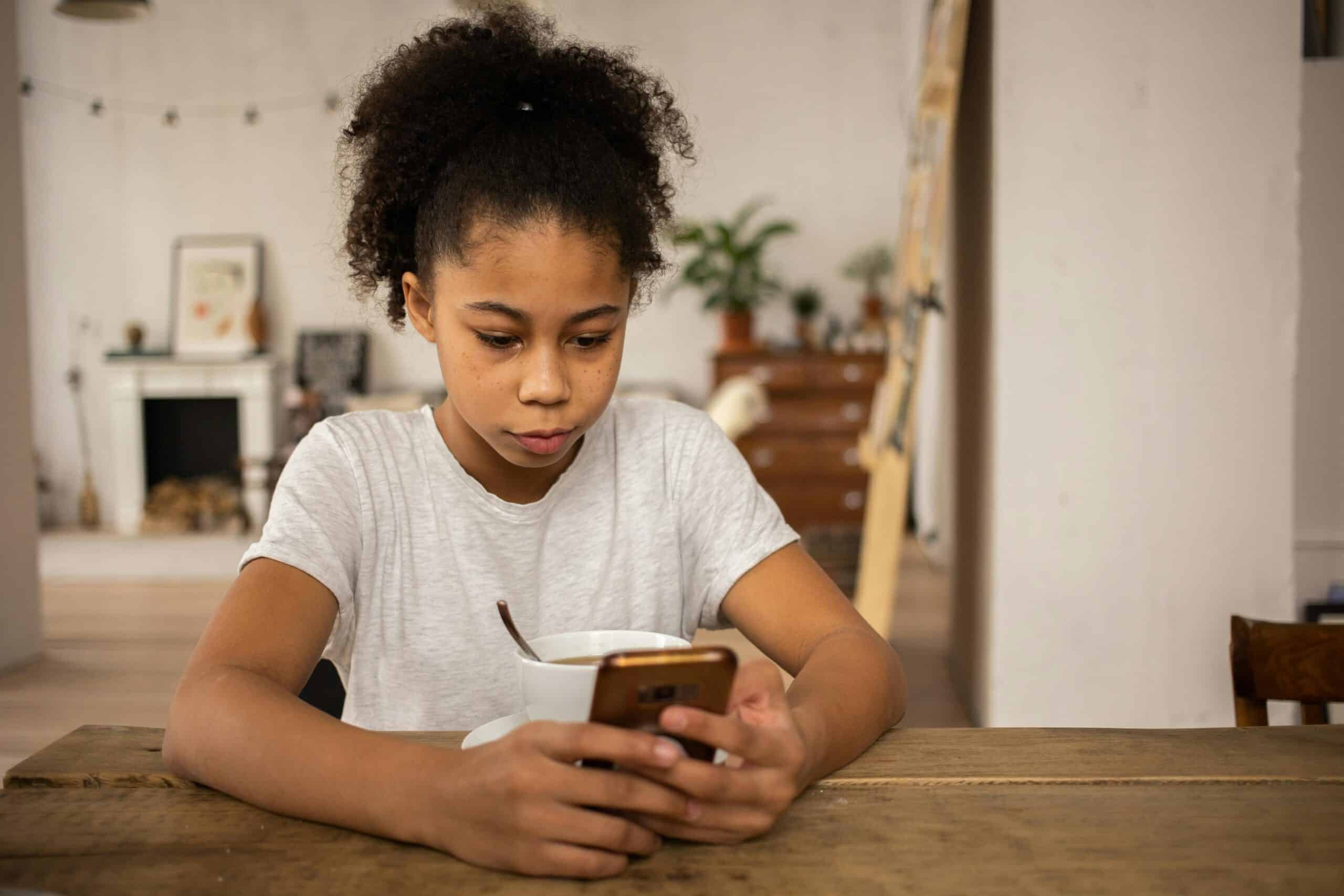 Young girl - under 16 years of age - in a white t shirt sitting at table using mobile phone.