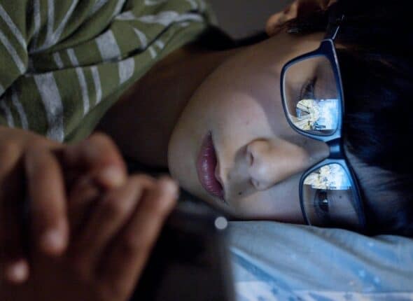 Young boy wearing black framed eyeglasses lying in bed looking at his phone in the dark.