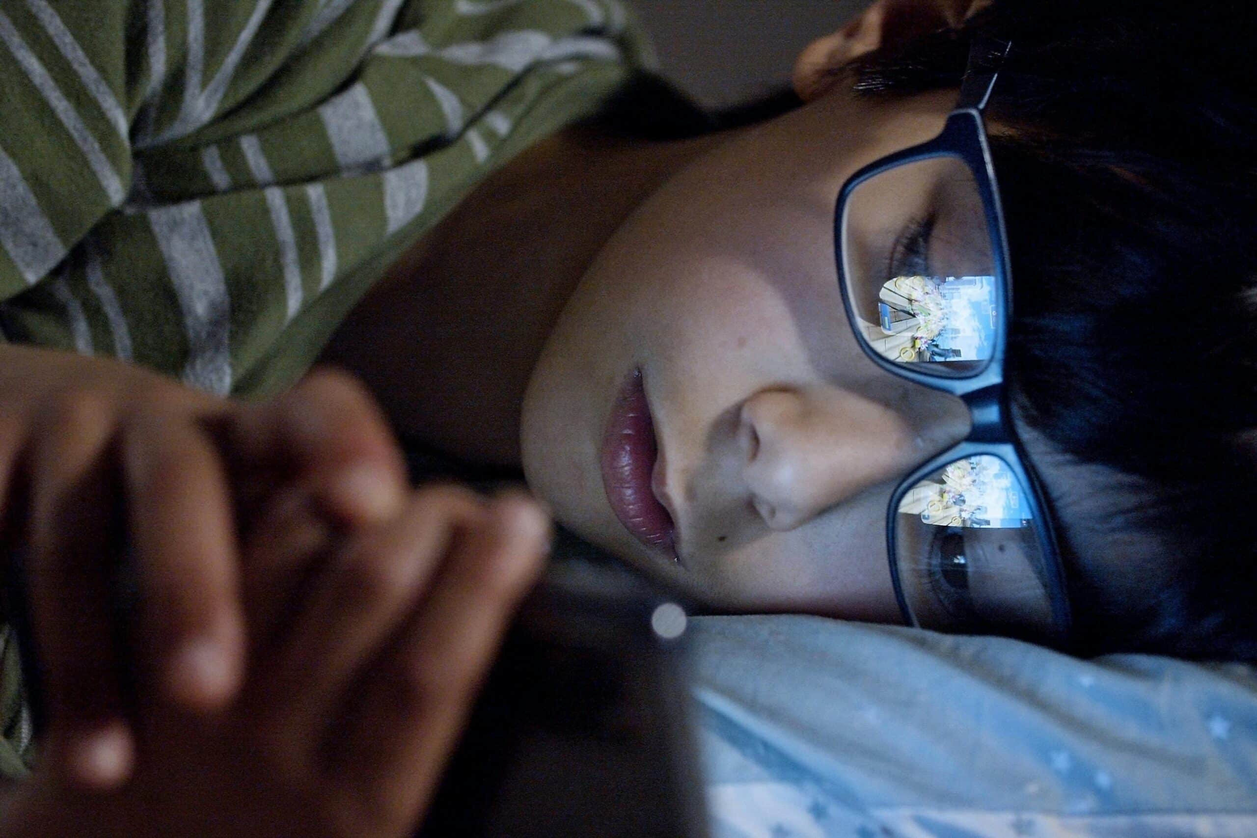 Young boy wearing black framed eyeglasses lying in bed looking at his phone in the dark.