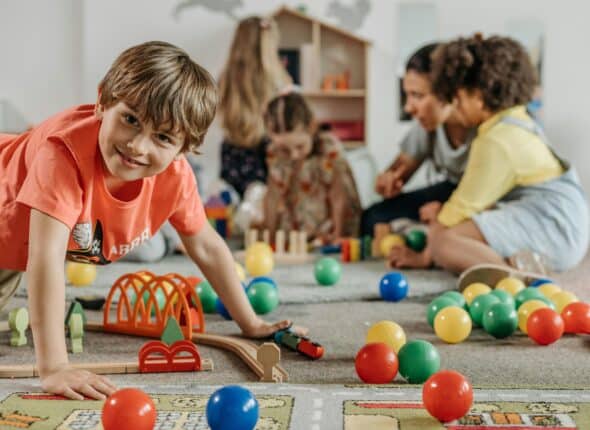 Smiling young boy playing on the floor with toys and in the background an adult it playing with other kids.