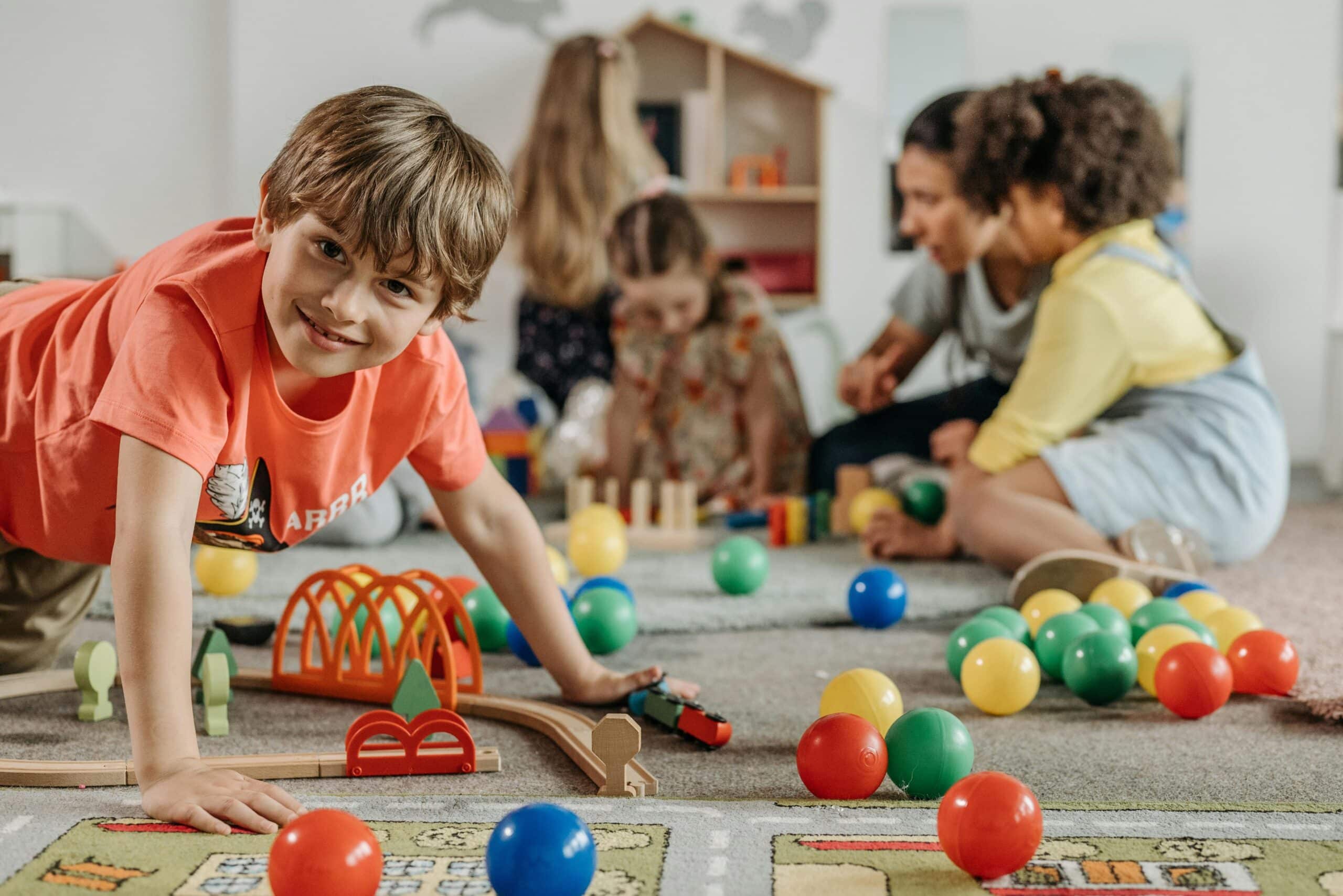 Smiling young boy playing on the floor with toys and in the background an adult it playing with other kids.