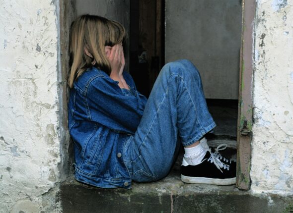 Lonely girl wearing denim sits in a door covering her face with her hands.