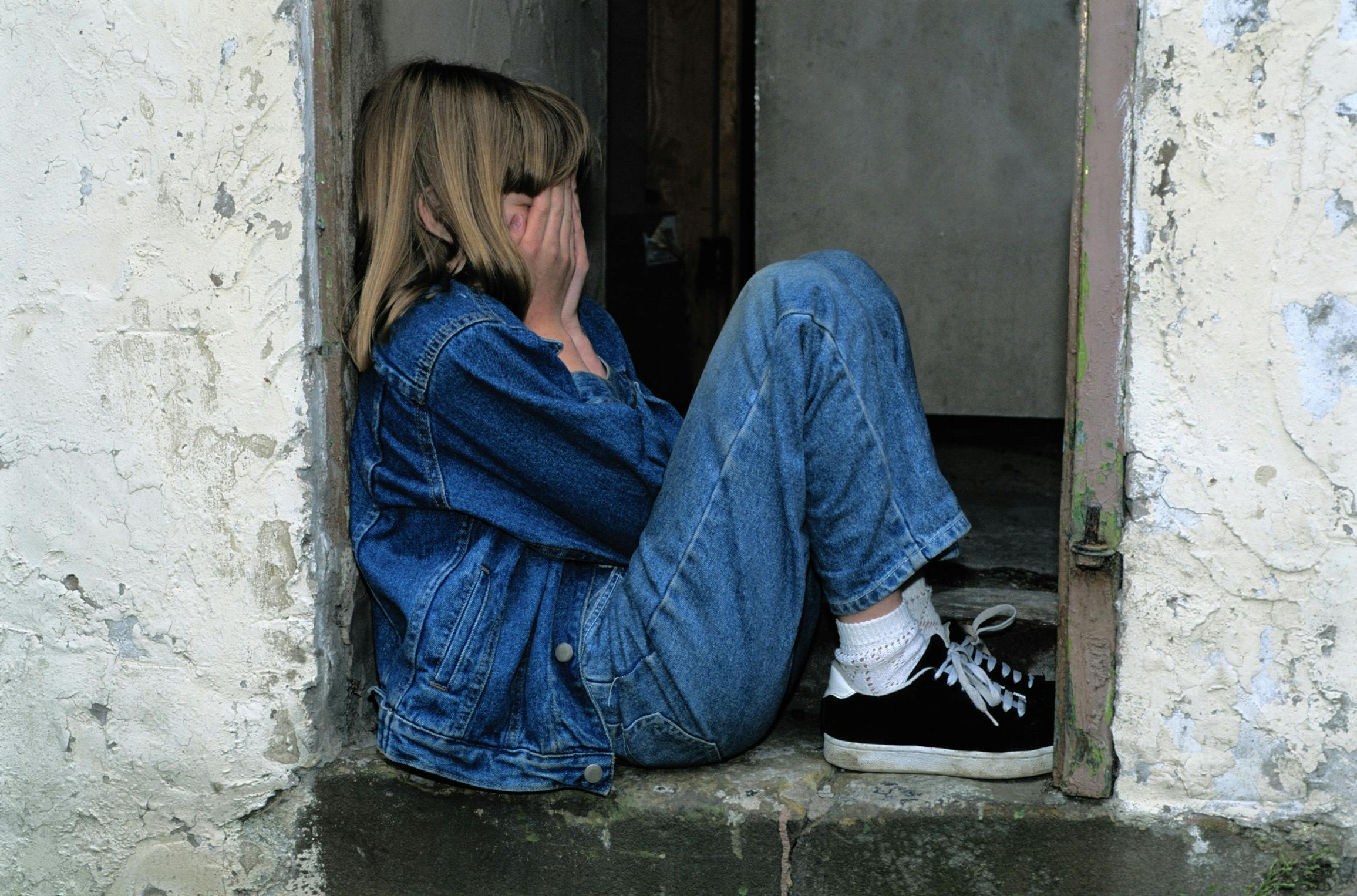Lonely girl wearing denim sits in a door covering her face with her hands.