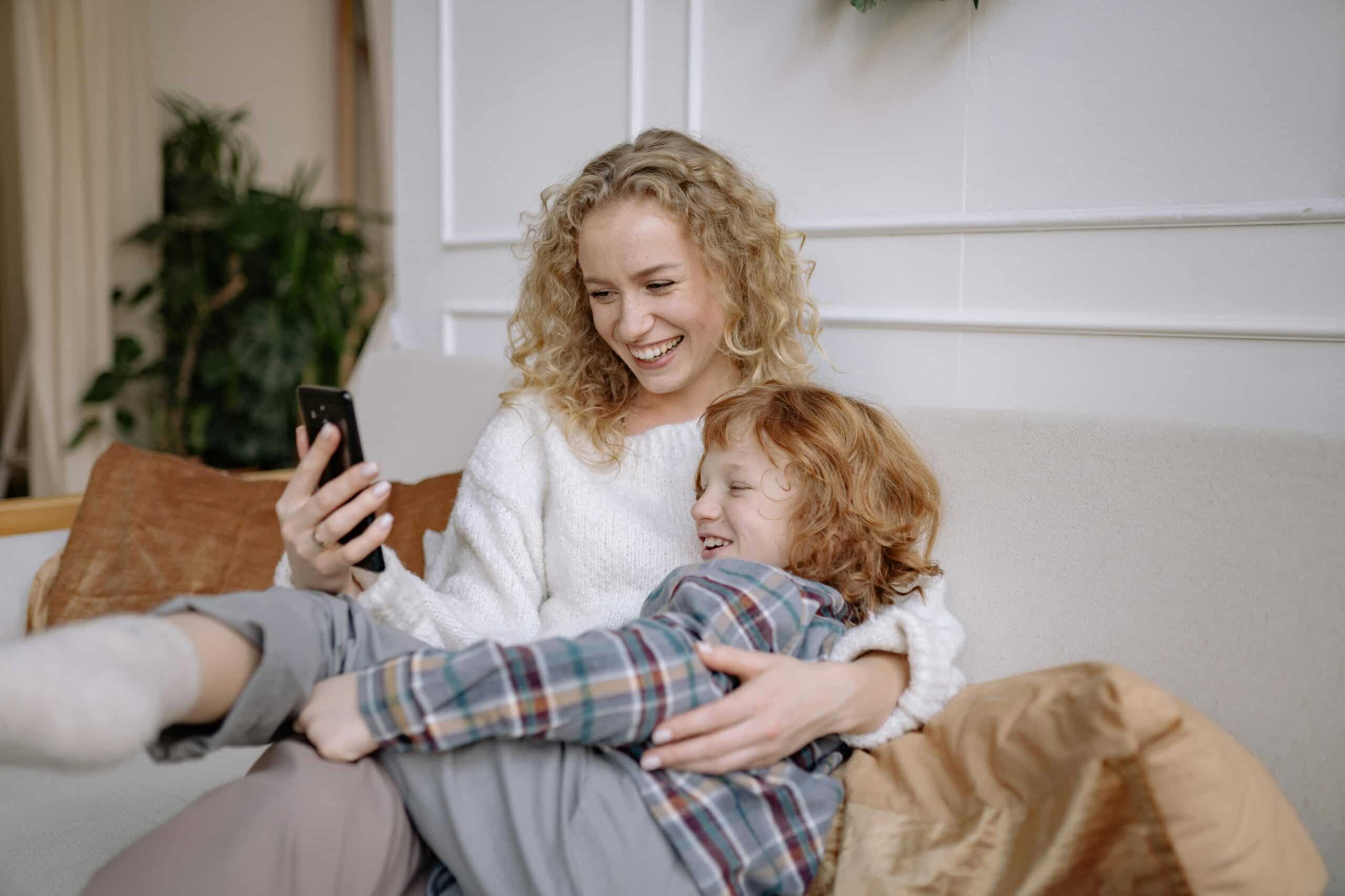 Mother and son sitting on a sofa smiling and looking at a mobile phone.