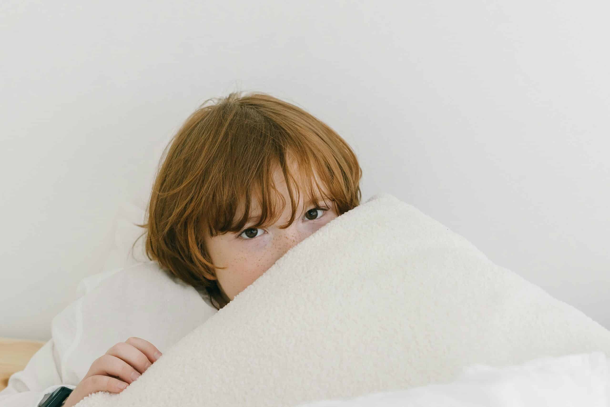Shy red headed boy peeking out from under a white blanket.