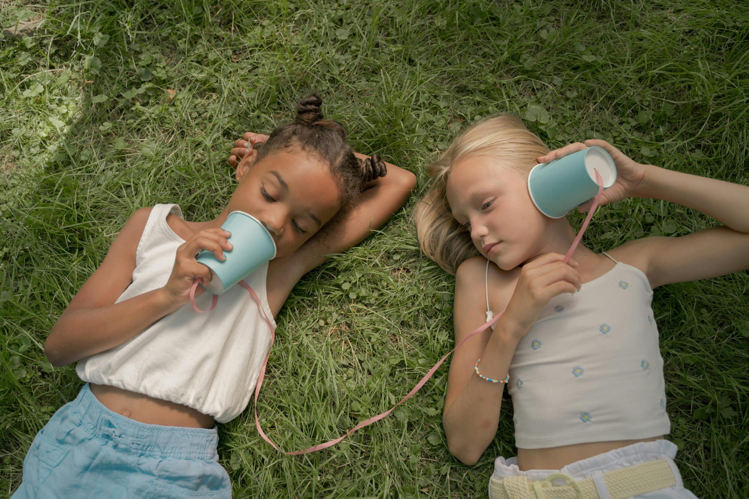 Two young girls laying on the grass playing telephone calls using paper paper cups on a string.